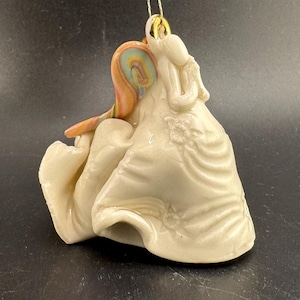 May include: A cream-coloured ceramic ornament depicting two figures embracing. One figure has a swirl of orange, yellow, and blue on its head. The ornament has a gold string for hanging. The figures are dressed in flowing robes with floral details.