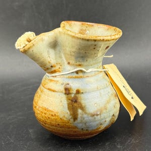 Könnte beinhalten: Handgefertigter Keramikkrug mit einem einzigartigen Ausguss und einem abgerundeten Körper. Der Krug hat eine Mischung aus Erdtönen, darunter Braun, Blau und Beige, mit einer strukturierten Oberfläche. Ein kleines Etikett ist mit einer Schnur befestigt. Der Krug ist etwa 13 cm hoch.