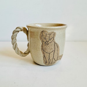 May include: A beige ceramic mug with a rope-like handle and a black line drawing of a cat on the front.