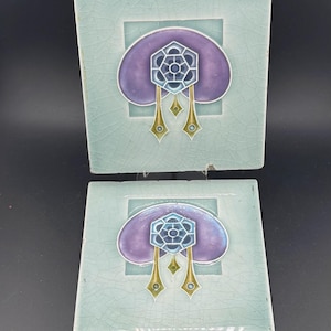 Puede incluir: Dos baldosas de cerámica cuadradas con un fondo verde azulado claro y un diseño central. El diseño presenta una forma púrpura en forma de nube con una pieza central geométrica azul y plateada y acentos dorados colgantes. Las baldosas tienen un esmalte craquelado.