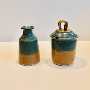 May include: A set of two ceramic containers with a blue and brown glaze. The smaller container is a vase with a narrow neck and a wide base. The larger container has a lid with a loop handle.