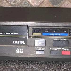 Puede incluir: Un reproductor de audio digital de disco compacto negro con el nombre de la marca "MultiTech" y el número de modelo "MD-150" impresos en la parte delantera. El reproductor tiene una pantalla digital que muestra "8.8.88" y botones para reproducir, pausar, detener, avanzar, retroceder y abrir/cerrar.