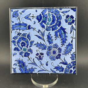 Può includere: Piastrella quadrata in ceramica con un motivo floreale nei toni del blu. La piastrella presenta uno sfondo azzurro chiaro con motivi floreali blu scuro e un bordo nero. La piastrella è esposta su un supporto trasparente.