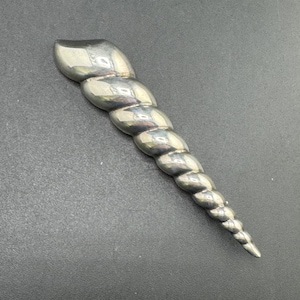 Può includere: Un oggetto in metallo color argento lucido a forma di corno conico e a spirale. La superficie è liscia e riflettente, caratterizzata da una serie di creste curve e segmentate che diminuiscono gradualmente di dimensioni dalla base alla punta.