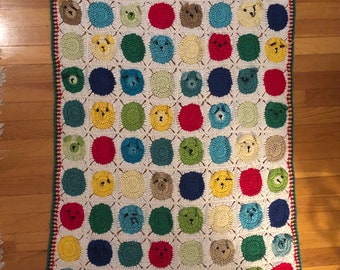 May include: A crocheted blanket with a repeating pattern of colorful circles featuring animal faces. The blanket has a white background with green, red, blue, and yellow circles.