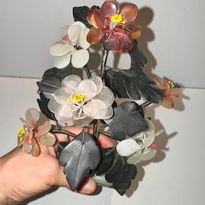 May include: A decorative floral sculpture featuring flowers in shades of white, brown, and translucent colors. The flowers have yellow and red accents, with dark gray leaves and stems. The sculpture is held in a hand.