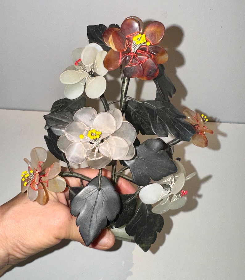 May include: A decorative floral sculpture featuring flowers in shades of white, brown, and translucent colors. The flowers have yellow and red accents, with dark gray leaves and stems. The sculpture is held in a hand.