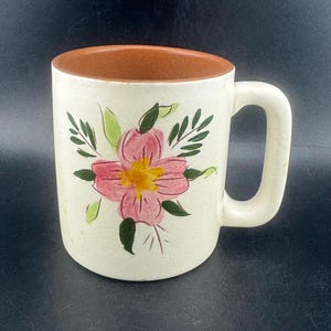 Może przedstawiać: Kremowy ceramiczny kubek z kwiatowym wzorem. Kubek ma różowy kwiat z żółtym środkiem i zielonymi liśćmi. Wewnętrzna krawędź jest brązowa, a uchwyt biały.