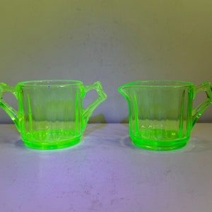 May include: Two vintage green glass sugar and creamer set. The creamer has a handle on each side and the sugar bowl has a single handle.