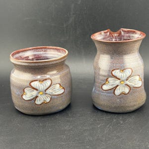 May include: Two handmade ceramic pieces. One is a small, round jar, and the other is a taller, vase-like container. Both are a mottled gray-brown with a white and yellow flower design. The rims are a deep burgundy color.