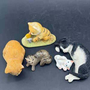 May include: A selection of ceramic cat ornaments in different poses and colours. Includes a tabby kitten, a black and white cat, and a ginger cat. The ornaments are detailed and appear to be hand-painted.