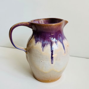 Könnte beinhalten: Ein kleiner Keramik-Krug mit einem hellbraunen Boden und einer weißen Glasur. Der Krug hat eine violette Glasur, die an der Seite herunterläuft und ein einzigartiges und künstlerisches Aussehen erzeugt.