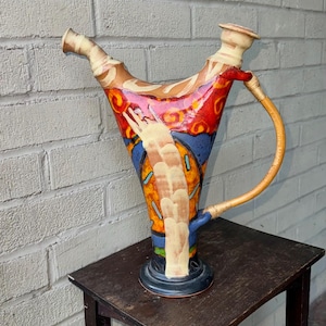May include: A colorful ceramic pitcher with a unique, abstract design. The pitcher has a wide, flared spout and a handle made of woven material. The pitcher is decorated with red, orange, yellow, blue, and white paint.