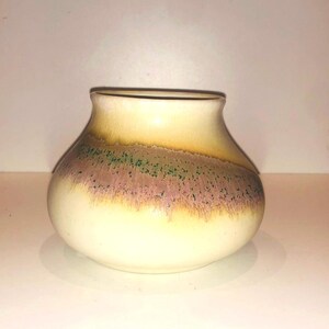 Puede incluir: Un jarrón pequeño y redondo de cerámica con un esmalte blanco y una banda de marrón, rosa y verde alrededor del centro.