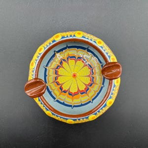 May include: A round ceramic ashtray with a blue and yellow floral design. The ashtray has two brown side compartments for cigarettes.