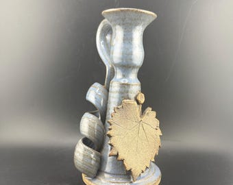 Könnte beinhalten: Ein Kerzenhalter aus Keramik in hellblauer Glasur mit braunem Sockel. Der Halter hat einen Griff, dekorative Wirbel und ein strukturiertes Blattdesign. Ein einzigartiges Stück für die Inneneinrichtung.