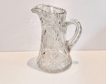 May include: Clear glass pitcher with a decorative floral and geometric pattern. The pitcher has a handle and a spout for pouring. The glass is clear, allowing light to pass through, and is suitable for serving beverages.