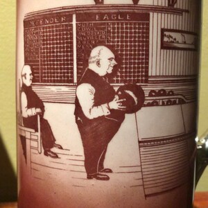 May include: A brown ceramic mug featuring a red and white illustration of two men bowling. The illustration includes a scoreboard with the words "Defender" and "Eagle" and the names of players. The mug is in a vintage style.