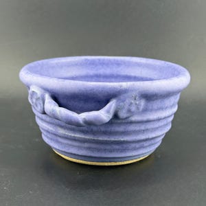 May include: A handmade, lavender-colored ceramic bowl with a textured, ribbed design. The bowl features two rope-like handles and a light tan base. The bowl is a unique piece of pottery.