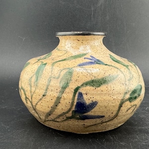 May include: A speckled, beige ceramic vase with a rounded body and a narrow neck. The vase is decorated with blue flower and green leaf designs. The rim is painted black. A decorative piece for the home.