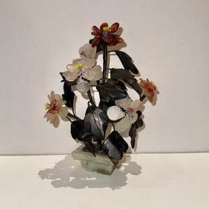 May include: A decorative floral arrangement in a square, light green stone pot. The arrangement features dark leaves and flowers in shades of white, red, and brown. The flowers have yellow centers. The piece is set against a white background.