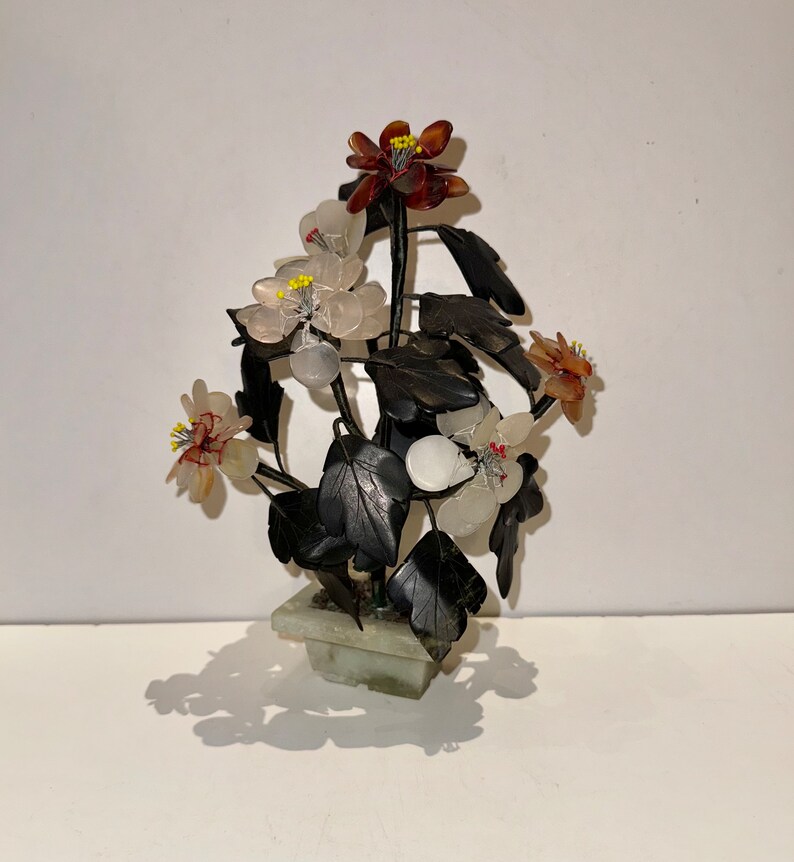 May include: A decorative floral arrangement in a square, light green stone pot. The arrangement features dark leaves and flowers in shades of white, red, and brown. The flowers have yellow centers. The piece is set against a white background.