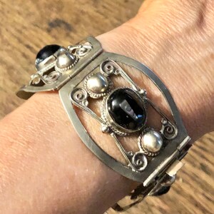 Puede incluir: Brazalete de pu&ntilde;o plateado con un dise&ntilde;o ornamentado. El brazalete presenta tres piedras negras grandes de forma ovalada y varios acentos plateados m&aacute;s peque&ntilde;os. El brazalete se asegura con un cierre.