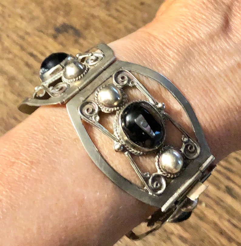 Puede incluir: Brazalete de pu&ntilde;o plateado con un dise&ntilde;o ornamentado. El brazalete presenta tres piedras negras grandes de forma ovalada y varios acentos plateados m&aacute;s peque&ntilde;os. El brazalete se asegura con un cierre.