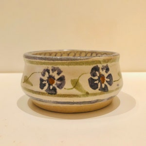 Puede incluir: Un pequeño cuenco de cerámica blanca con un diseño floral verde y azul. El cuenco tiene una forma redondeada y un borde ligeramente acampanado.