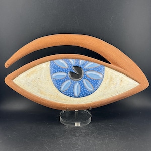 May include: A ceramic wall sculpture shaped like an eye with a terracotta-colored frame. The iris is a vibrant blue with a pattern of light blue petals and a black pupil. The sclera is an off-white speckled glaze. It rests on a clear acrylic stand.