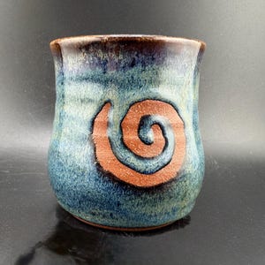 May include: Handmade ceramic mug with a blue and green glaze. A large, reddish-brown spiral design is centered on the mug. The rim of the mug is a darker brown. The mug is a unique piece of pottery.