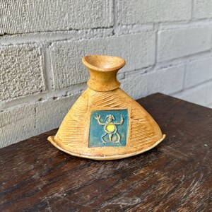 May include: A small, ceramic vase with a triangular base and a narrow neck. The vase is decorated with a stylized, blue and yellow creature in a square frame.