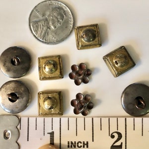 May include: A selection of vintage buttons in a variety of shapes, sizes and colours, including silver, gold and brown. The buttons are displayed on a white surface with a ruler for scale. The ruler is marked in inches.