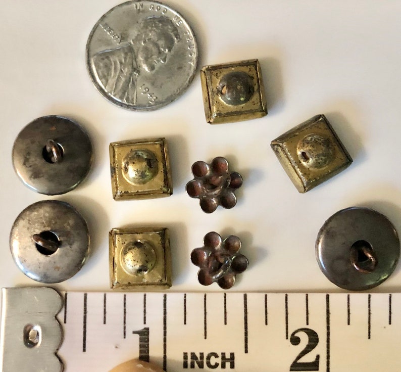 May include: A selection of vintage buttons in a variety of shapes, sizes and colours, including silver, gold and brown. The buttons are displayed on a white surface with a ruler for scale. The ruler is marked in inches.