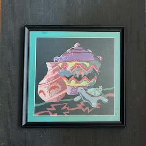 May include: A framed cross-stitch artwork depicting a still life of three colorful ceramic pots and a turtle. The pots are decorated with geometric patterns in shades of purple, red, yellow, and green. The turtle is a light purple color with a green shell.