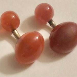 Puede incluir: Un par de gemelos con detalles de piedra redonda de color melocotón. Los gemelos están hechos de metal plateado.