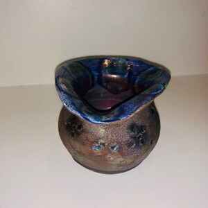 Puede incluir: Una pequeña maceta de cerámica hecha a mano con un esmalte azul y morado. La maceta tiene un cuerpo redondeado y una abertura ancha y acampanada. Está decorada con un patrón de pequeños puntos blancos.
