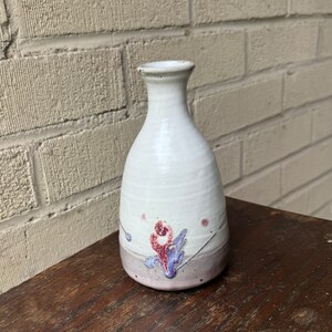 Pode incluir: Um pequeno vaso de cerâmica branca com um design floral rosa e roxo. O vaso tem um corpo arredondado e um pescoço estreito.