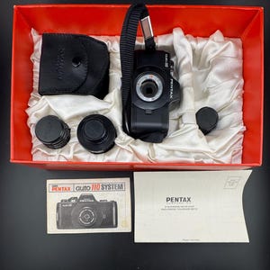 Puede incluir: Un kit de cámara Pentax Auto 110 negro en una caja roja, que incluye una cámara, lentes, una funda y documentos. La cámara tiene una correa negra y está rodeada de tela blanca. La caja también contiene un manual y un sobre de Pentax Corporation.
