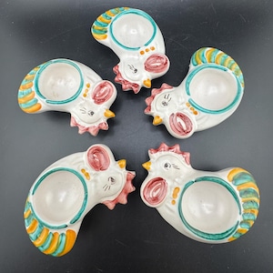 May include: Set of five ceramic egg cups shaped like chickens. Each cup is white with green, yellow, and red painted details. The chickens have a whimsical design with painted eyes and beaks.