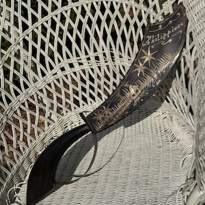 May include: A dark brown curved blade with a handle and a hand guard. The blade features an etched design of palm trees, a small house, and the word Philippines written in script. The surface of the blade has a polished finish with light details.