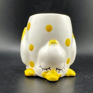 May include: A ceramic duck-shaped planter in white with yellow polka dots. The duck has a yellow beak and feet, and closed eyes. The planter is designed to look like a sleeping duck.