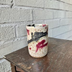 May include: A white ceramic mug with a speckled glaze and abstract black and pink designs.