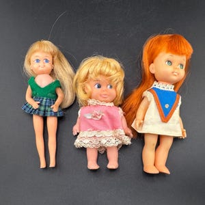 May include: Three vintage dolls with blonde and red hair. The dolls are wearing dresses in green, pink, and white with blue accents. The doll on the left has a green top and plaid skirt. The doll in the middle has a pink dress with lace trim.