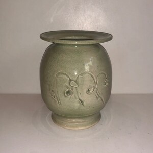 May include: A light green ceramic vase with a wide, round body and a flared rim. The vase is decorated with a raised design of stylized flowers.