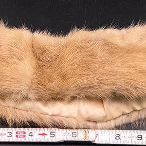 May include: A light brown fur headband, approximately 28 cm long, with a cream-coloured lining. The fur appears soft and plush, with a slightly curved shape. The headband is measured with a ruler.