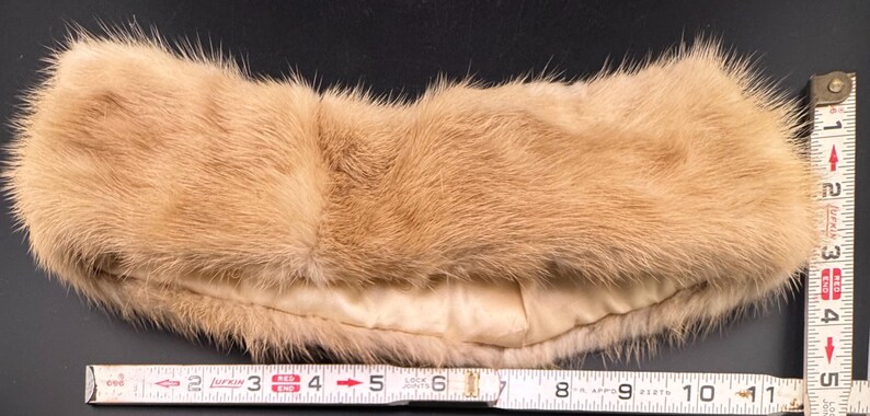 May include: A light brown fur headband, approximately 28 cm long, with a cream-coloured lining. The fur appears soft and plush, with a slightly curved shape. The headband is measured with a ruler.