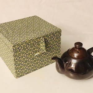 May include: A brown ceramic teapot with a lid and a spout, next to a square box with a green and blue geometric pattern.