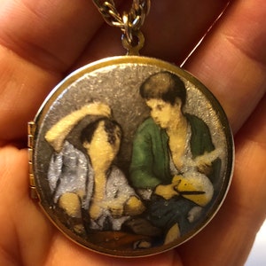 May include: A gold-toned locket pendant with a detailed miniature painting of two figures. The locket hangs from a gold-colored chain. The painting depicts two figures, one in a green jacket, the other in a light blue shirt.