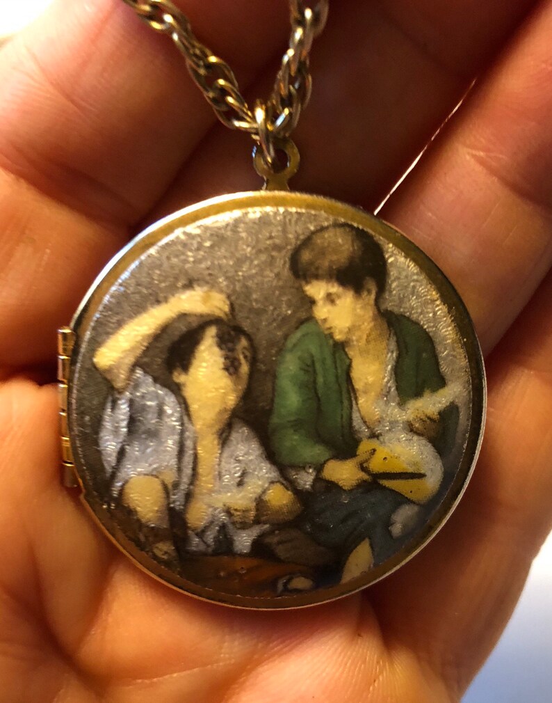 May include: A gold-toned locket pendant with a detailed miniature painting of two figures. The locket hangs from a gold-colored chain. The painting depicts two figures, one in a green jacket, the other in a light blue shirt.
